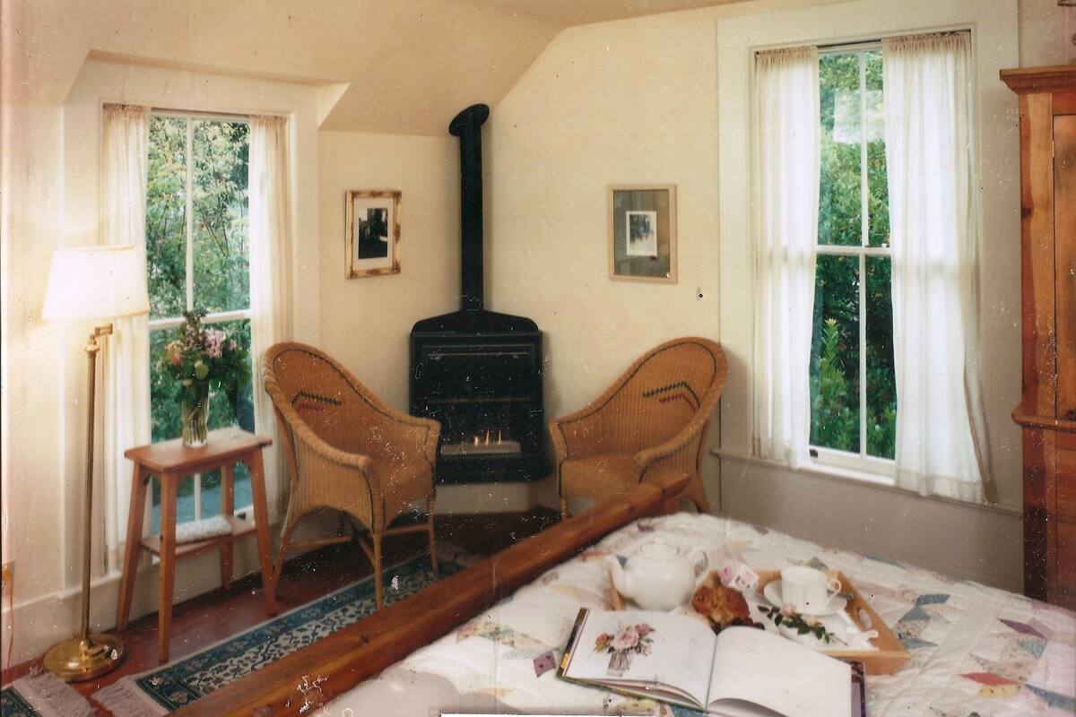 A room with a bed, two wicker chairs, a small table with a lamp, a fireplace, and two windows with curtains.