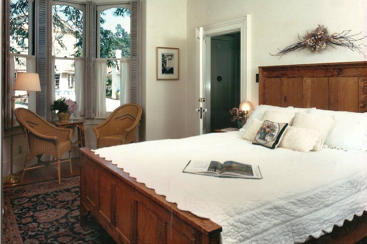 A bedroom with a wooden bed, a nightstand, a chair, a window with shutters, and a door.