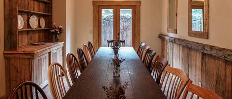 Une longue table à manger en bois, avec des chaises, un vase rempli de fleurs et des étagères en bois où sont disposées des assiettes.
