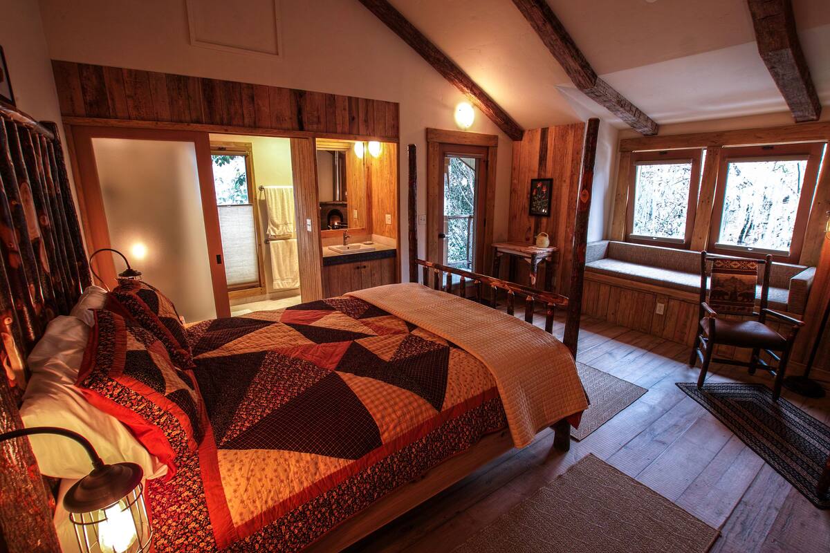 A bedroom with a large bed, a wooden ceiling, a window seat, and a bathroom with a mirror and sink.