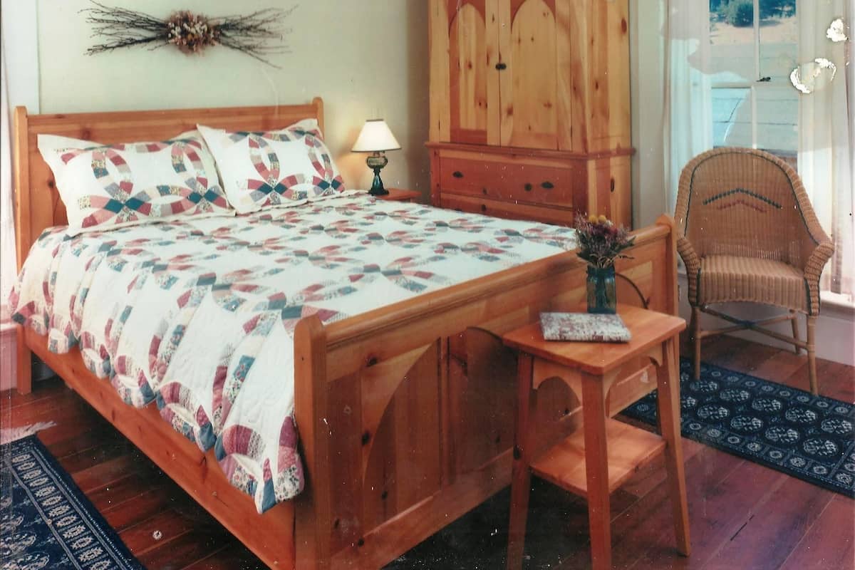 A bedroom with a wooden bed, a wooden nightstand, a wicker chair, a wooden cabinet, and a window with curtains.