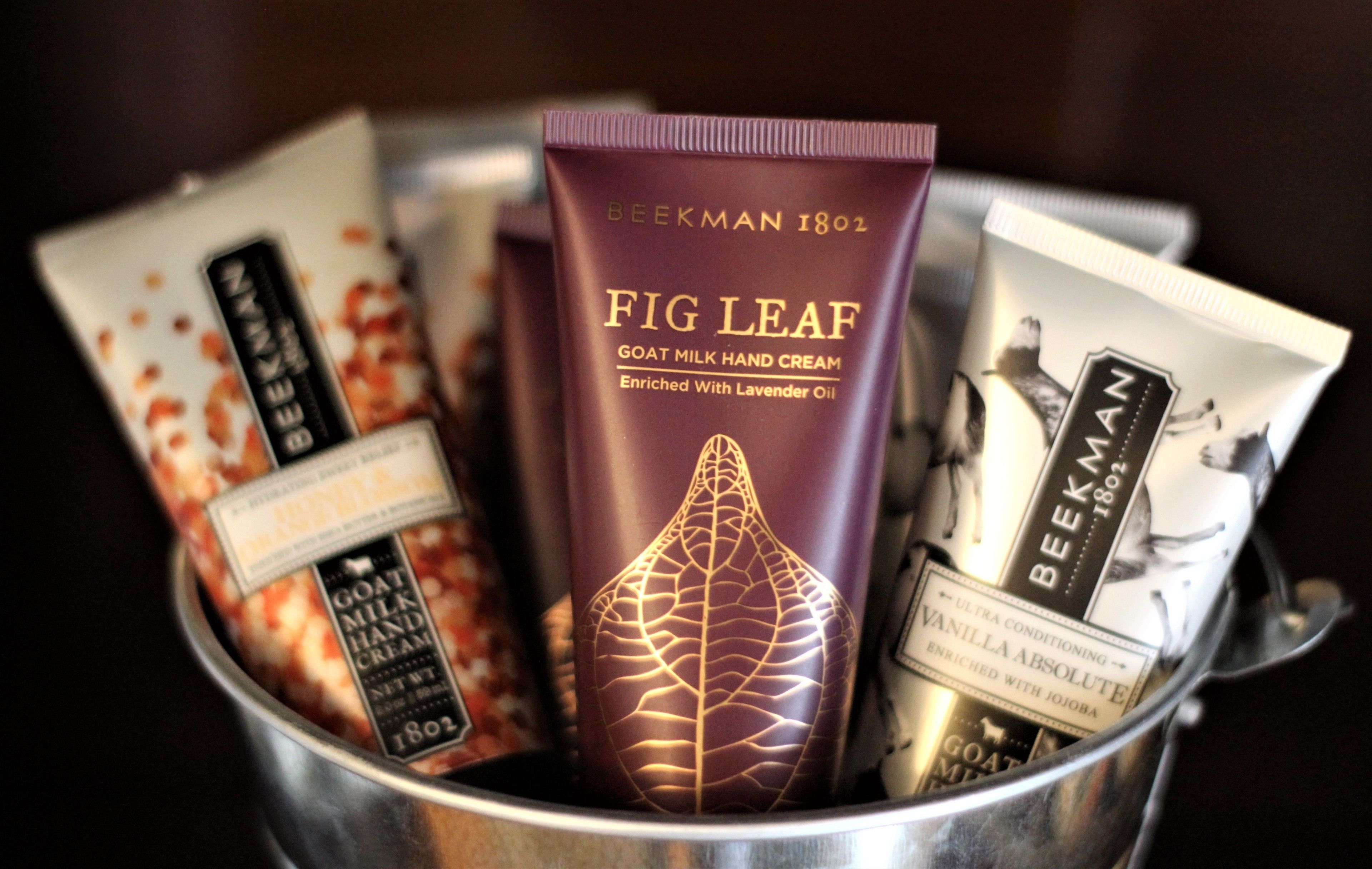 A metal container holding various skincare products, including a tube labeled "Fig Leaf Goat Milk Hand Cream."