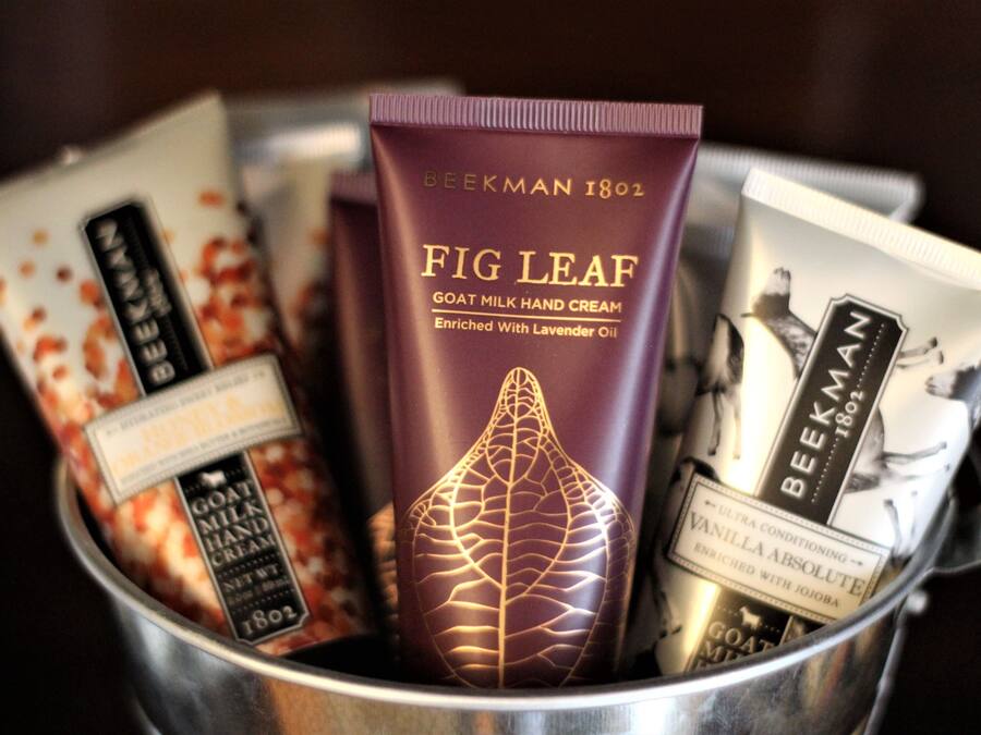 Recipiente de metal contendo diversos produtos de cuidados com a pele, incluindo um tubo com a etiqueta “Creme de Mãos de Figo e Leite de Cabra”.