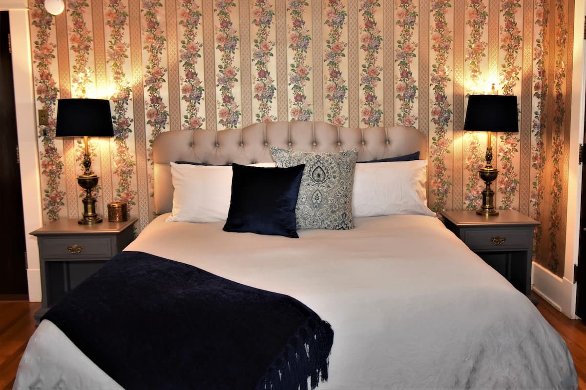 A bedroom with a tufted headboard, floral wallpaper, two bedside tables with lamps, and a dark throw blanket.