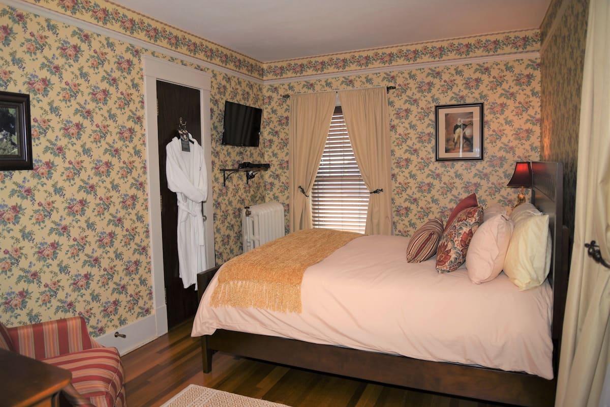 A bedroom with floral wallpaper, a bed with multiple pillows, a radiator, and a TV on the wall.