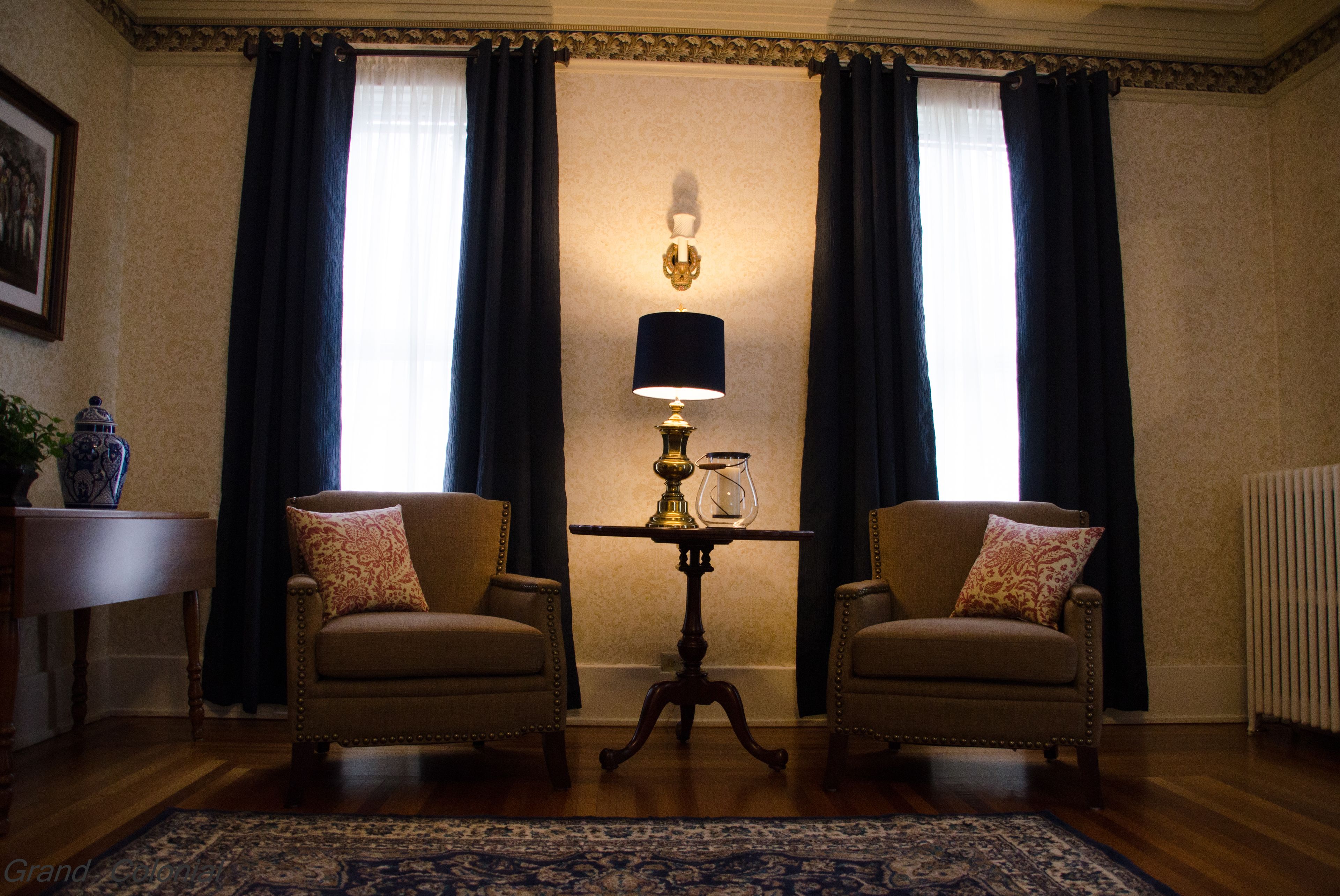 A room with two armchairs, a small table with a lamp, and a large window with curtains.