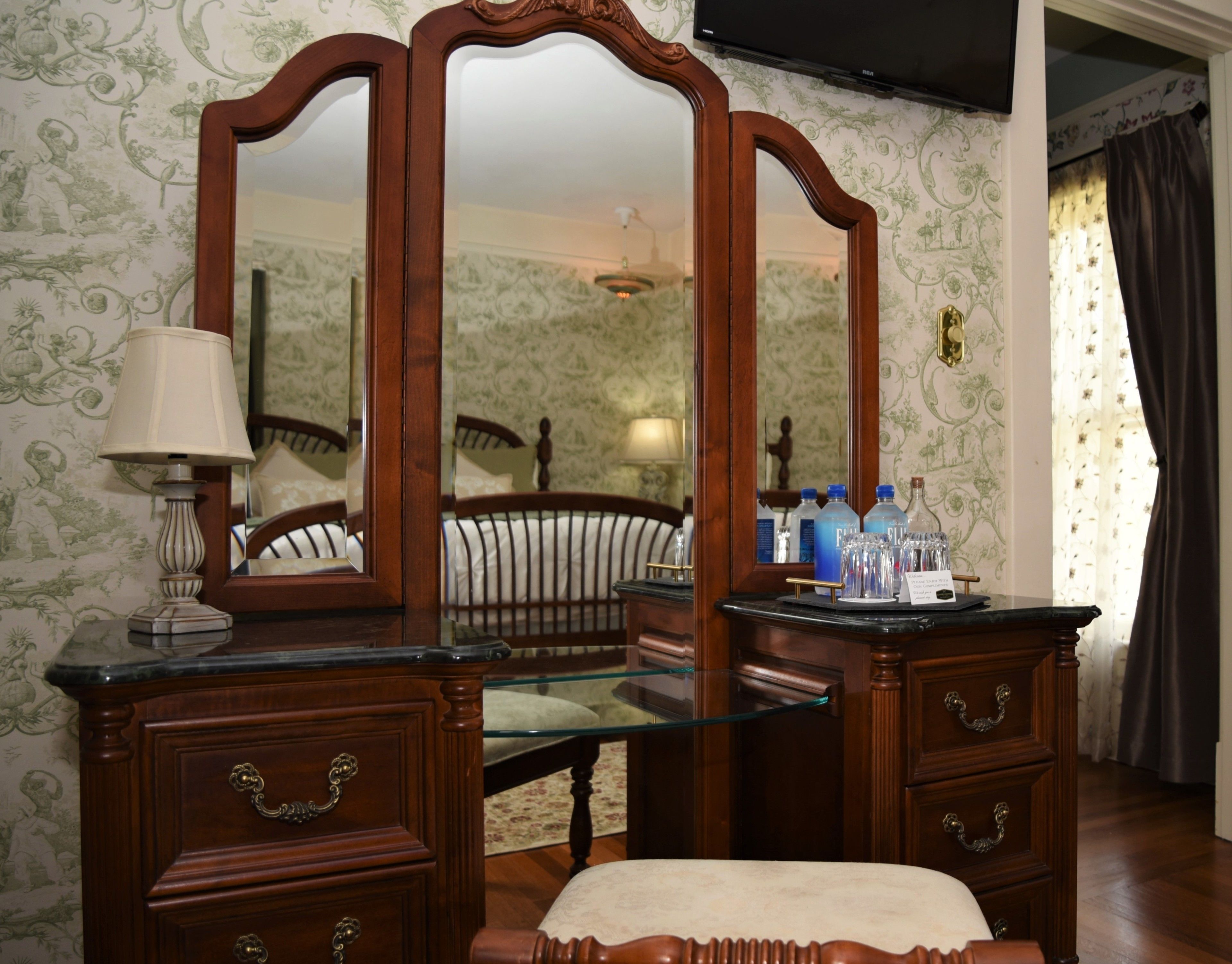 Una habitación con un tocador de madera, una mesa con cubierta de vidrio y un espejo. Hay dos cajones con tiradores de latón, una lámpara y una cama con cabecero de madera.