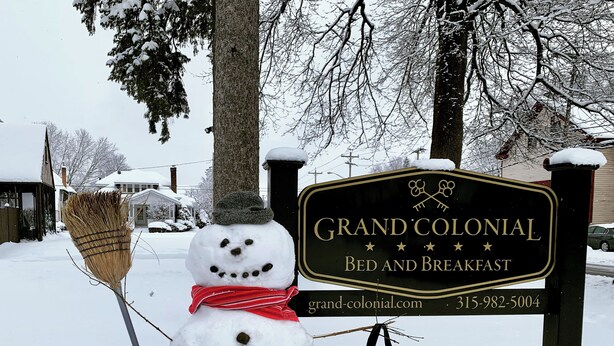 Un bonhomme de neige, tenant un balai et portant un sac, se trouve devant une enseigne indiquant « Grand Colonial Bed and Breakfast ».