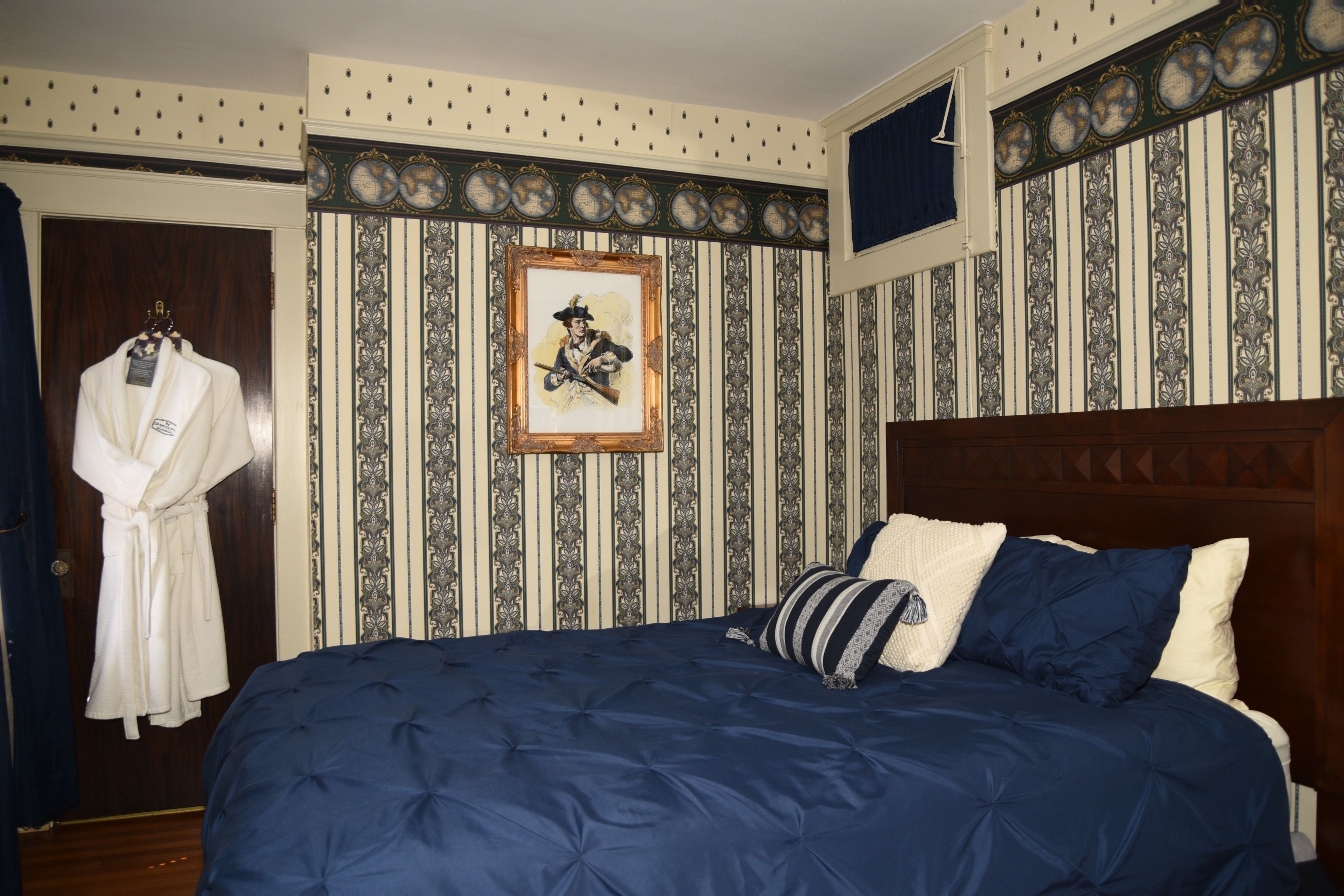 A bedroom with a large bed, striped wallpaper, a framed picture, and a robe hanging on the door.