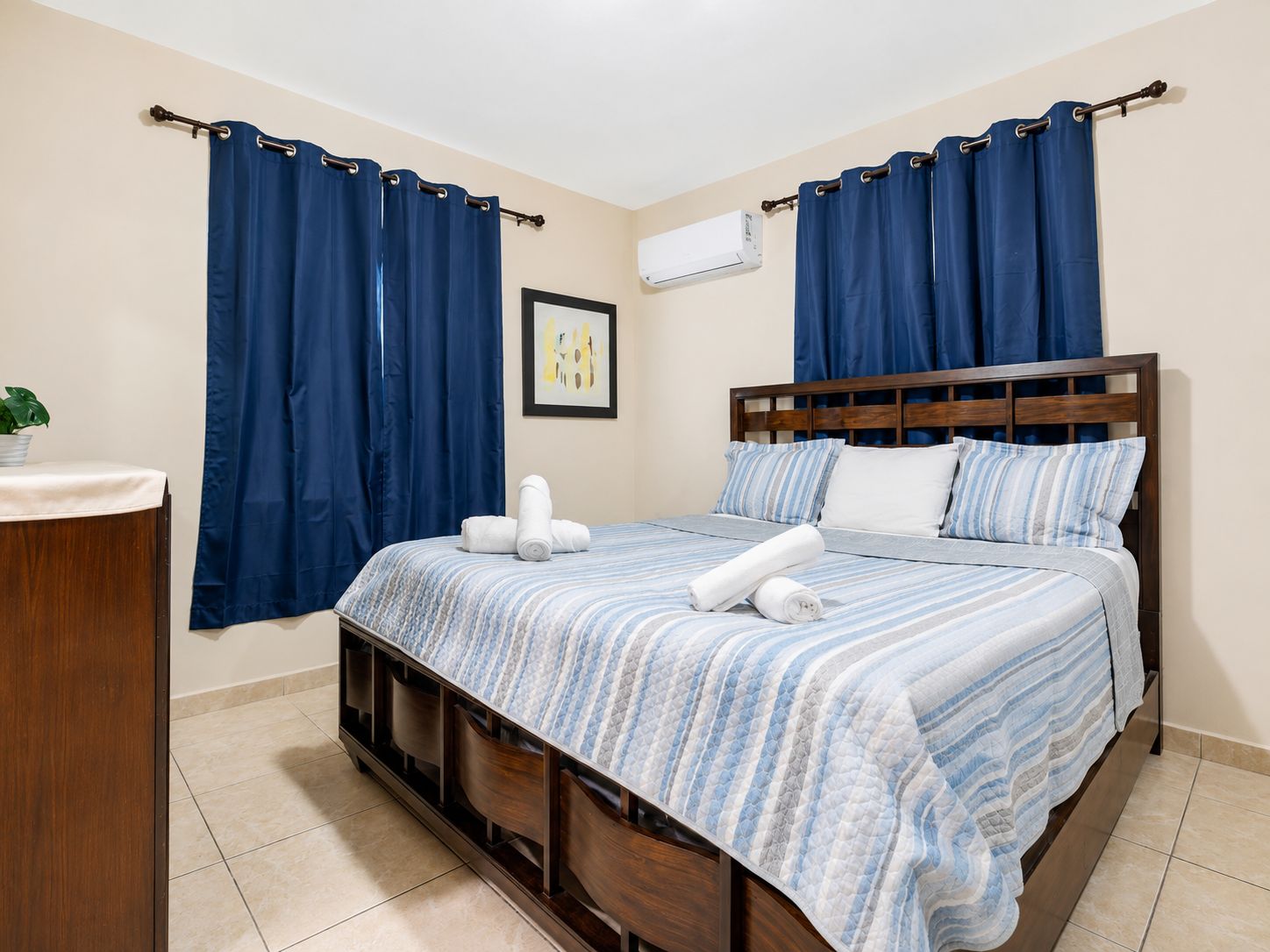 A hotel room with a bed, curtains, a bedside table, a plant, and an air conditioning unit.