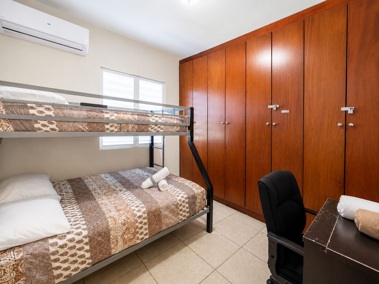 A bunk bed with patterned bedding, a desk with a chair, and wooden wardrobes.