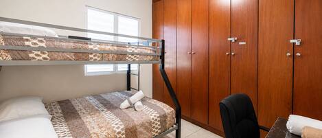 Een stapelbed met beddengoed met een patroon, een bureau met een stoel en houten kledingkasten.