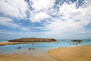 A beach with clear water, a sandy shore, and a rocky outcrop in the distance.
