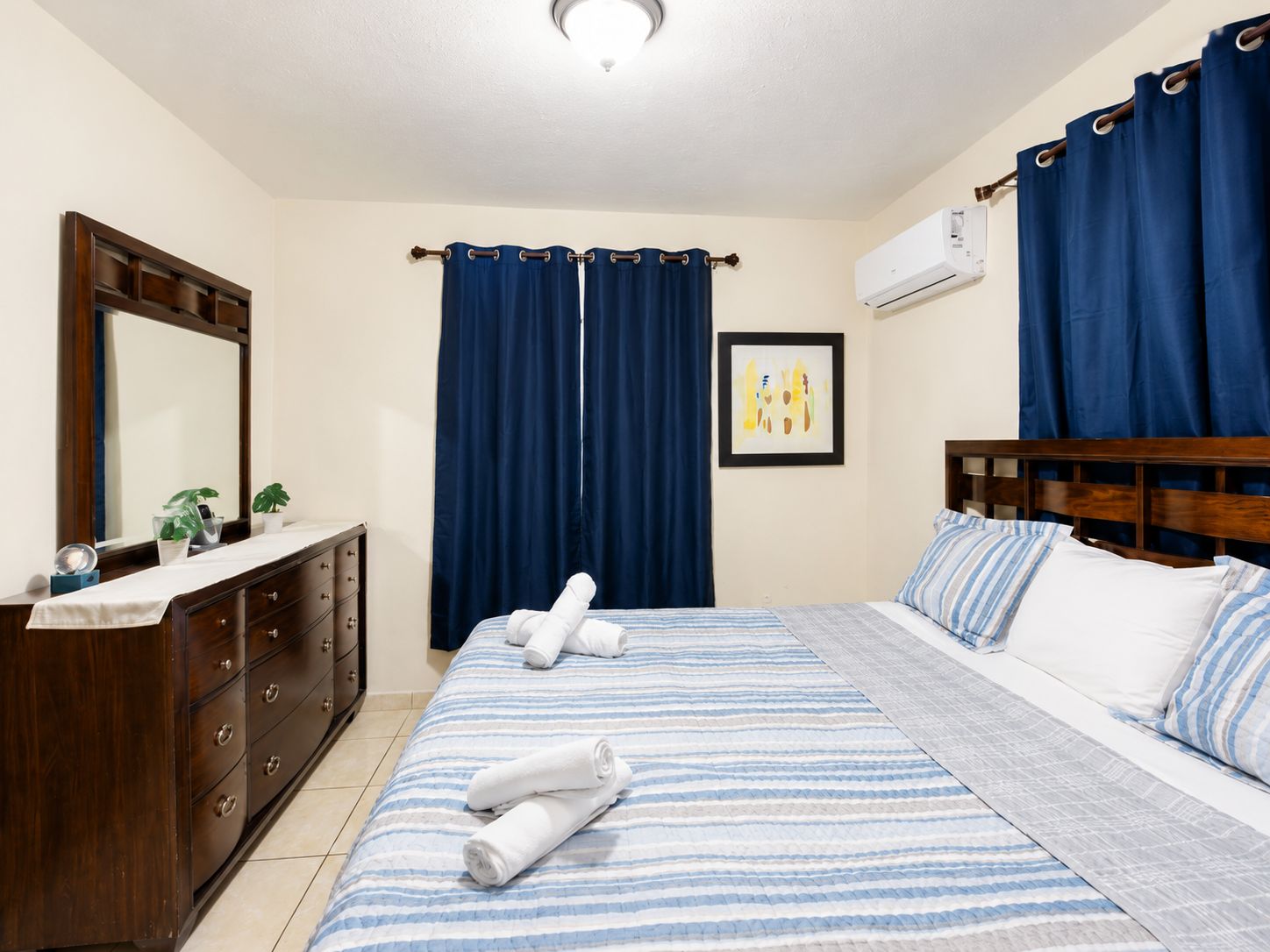 A hotel room with a bed, a dresser, a mirror, a painting, and blue curtains.