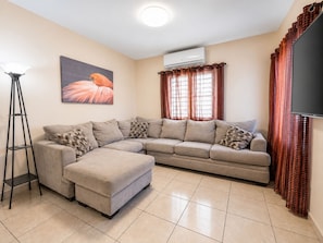 A living room with a sectional sofa, a standing lamp, an air conditioning unit, and a flat-screen TV. - Casa vacacional en Arenales Isabela (Isabela)