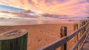 En strand med en träpromenad som leder ner till vattnet, en sandstrand och en himmel i solnedgångens f ärger.