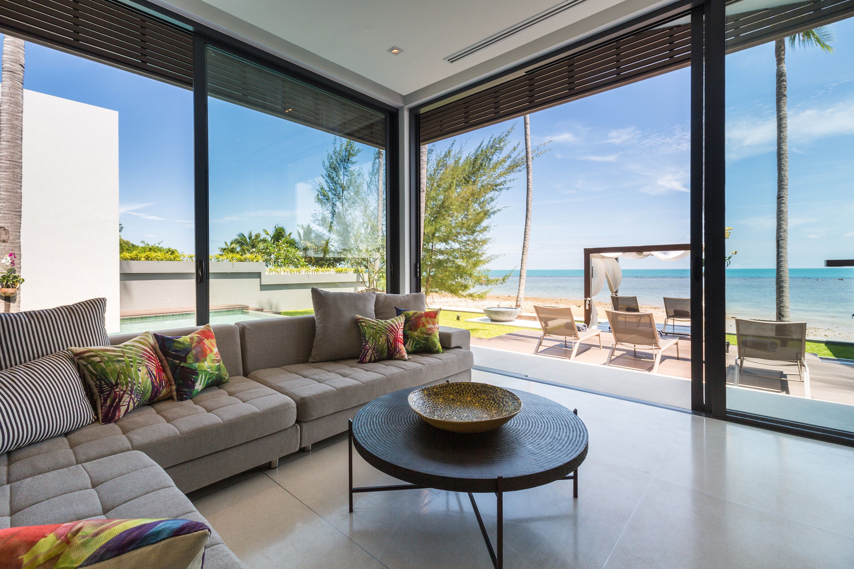 A modern living room with a large sofa, a coffee table, and a view of the beach and ocean through large windows.