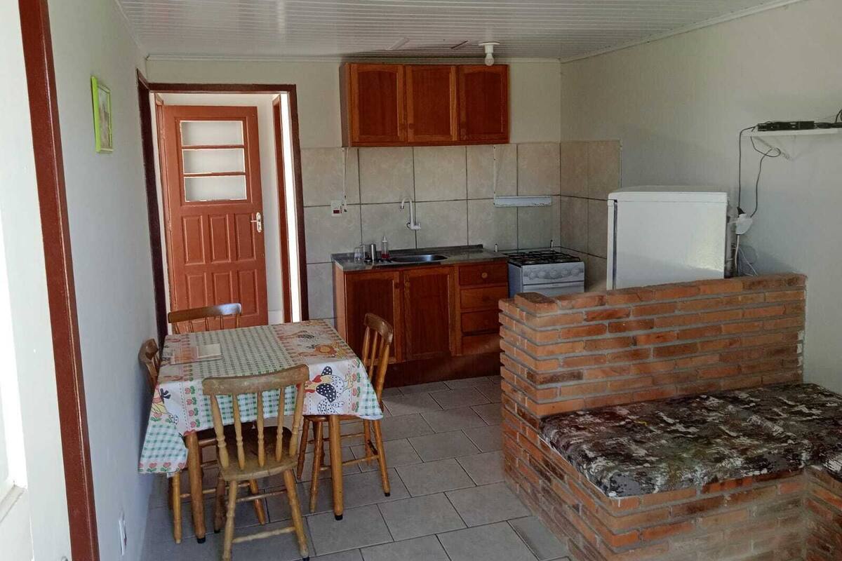 Cocina compacta con armarios de madera, estufa a gas, pileta y refrigerador. Hay una mesa de comedor con sillas y un pequeño banco de ladrillo.