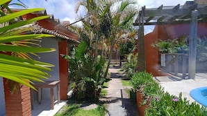 Un sentier bordé d’une végétation luxuriante et d’une piscine, qui mène à un bâtiment doté d’un toit en tuiles.