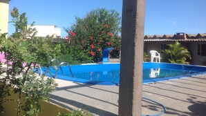 Une piscine dotée d’une plage bleue, entourée de verdure et bordée d’un bâtiment dont le toit est recouvert de tuiles.