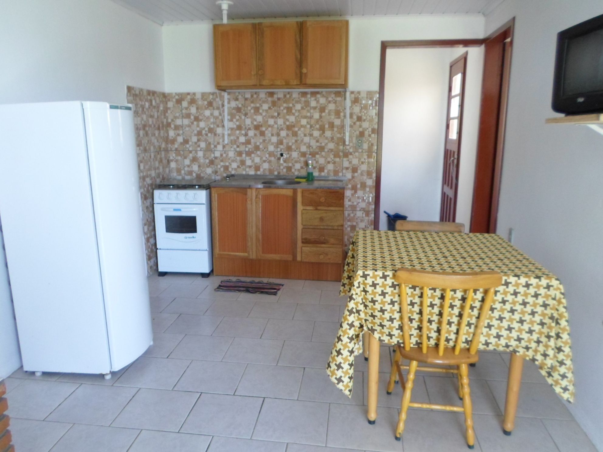 Cocina pequeña con refrigerador, estufa y armarios de madera. Una mesa de comedor con mantel estampado y sillas se encuentra cerca de una ventana.