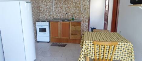 Cocina pequeña con refrigerador, estufa y armarios de madera. Una mesa de comedor con mantel estampado y sillas se encuentra cerca de una ventana.