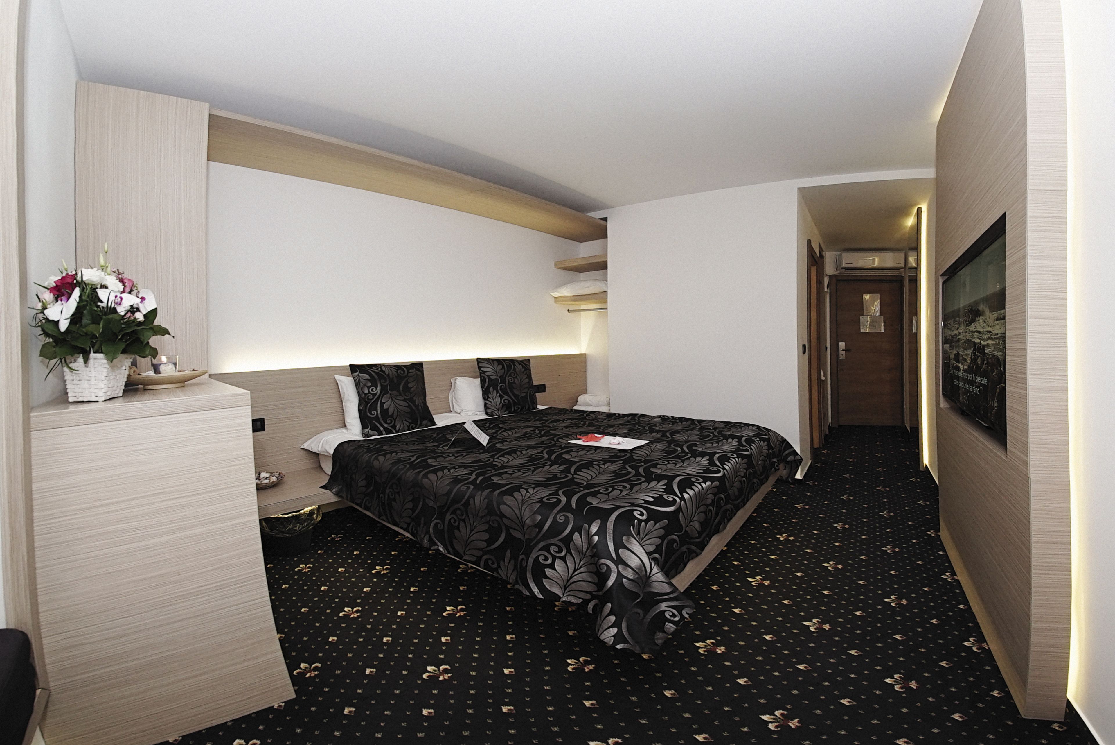 Habitación de hotel moderna con una cama grande, una mesita de noche con un florero y un televisor de pantalla plana montado en la pared, además de un pasillo que conduce a una puerta.