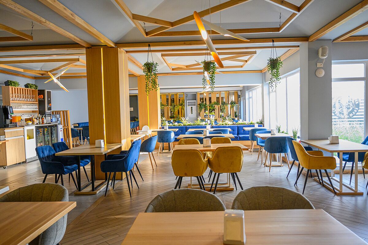 A modern restaurant with wooden beams, blue and yellow chairs, and hanging plants.