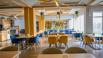 A modern restaurant with wooden beams, blue and yellow chairs, and hanging plants.