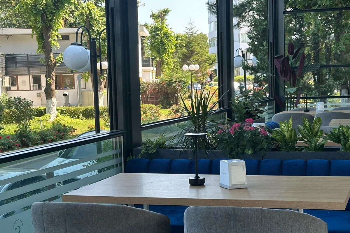 Een zitgedeelte binnen met uitzicht op een tuin en gebouwen. Er zijn blauwe, beklede bankjes, een tafel en een lamp.