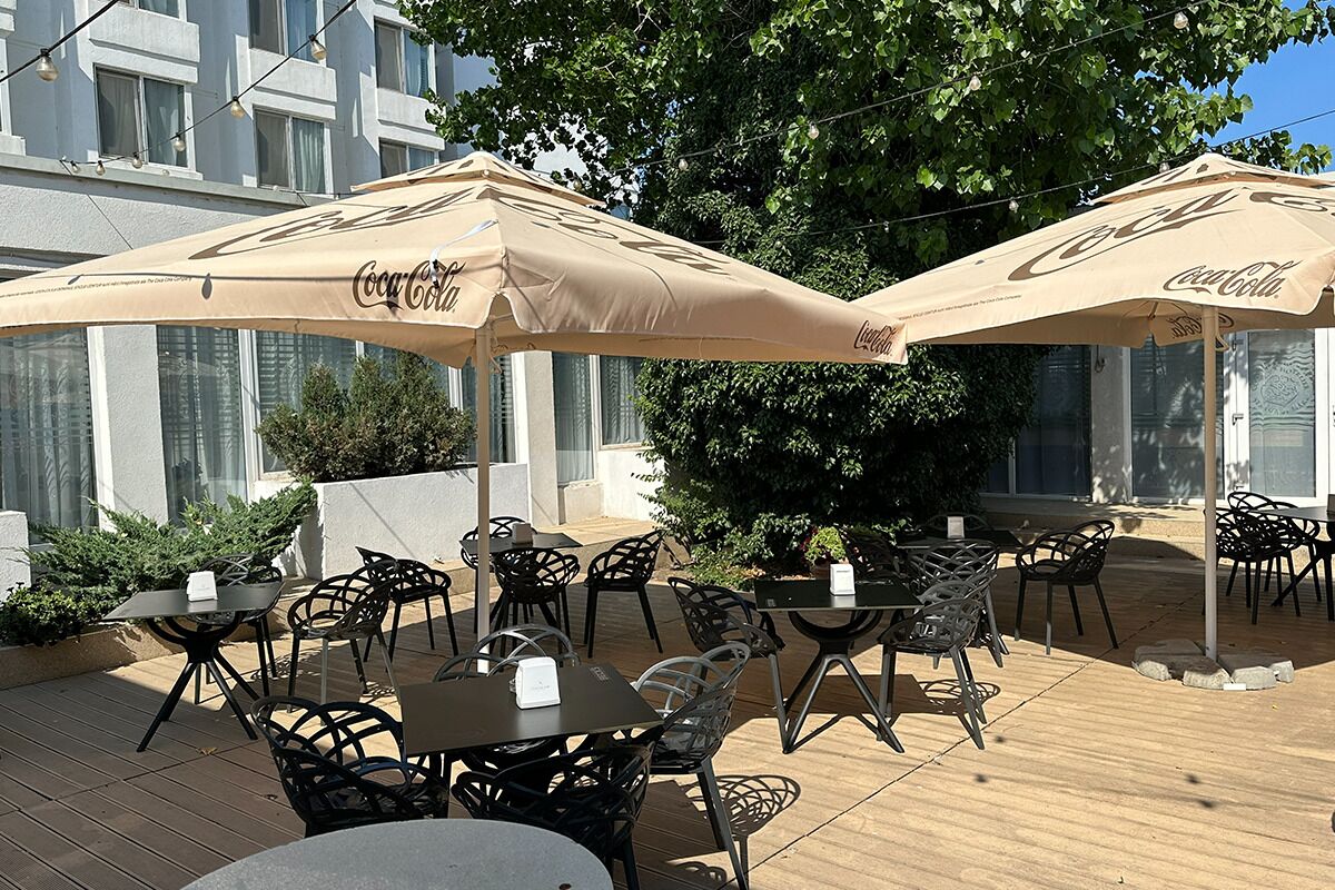 An outdoor seating area with tables and chairs under Coca-Cola branded umbrellas, surrounded by greenery and a building facade.