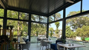 A covered outdoor seating area with tables and chairs, large windows, and potted plants. - Aqvatonic Balneo Spa - Steaua de Mare (Eforie)