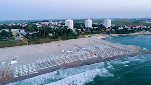 An aerial view of a beach with rows of sun loungers and umbrellas, a parking area, and a view of the sea and buildings in the background. - Aqvatonic Balneo Spa - Steaua de Mare (Eforie)