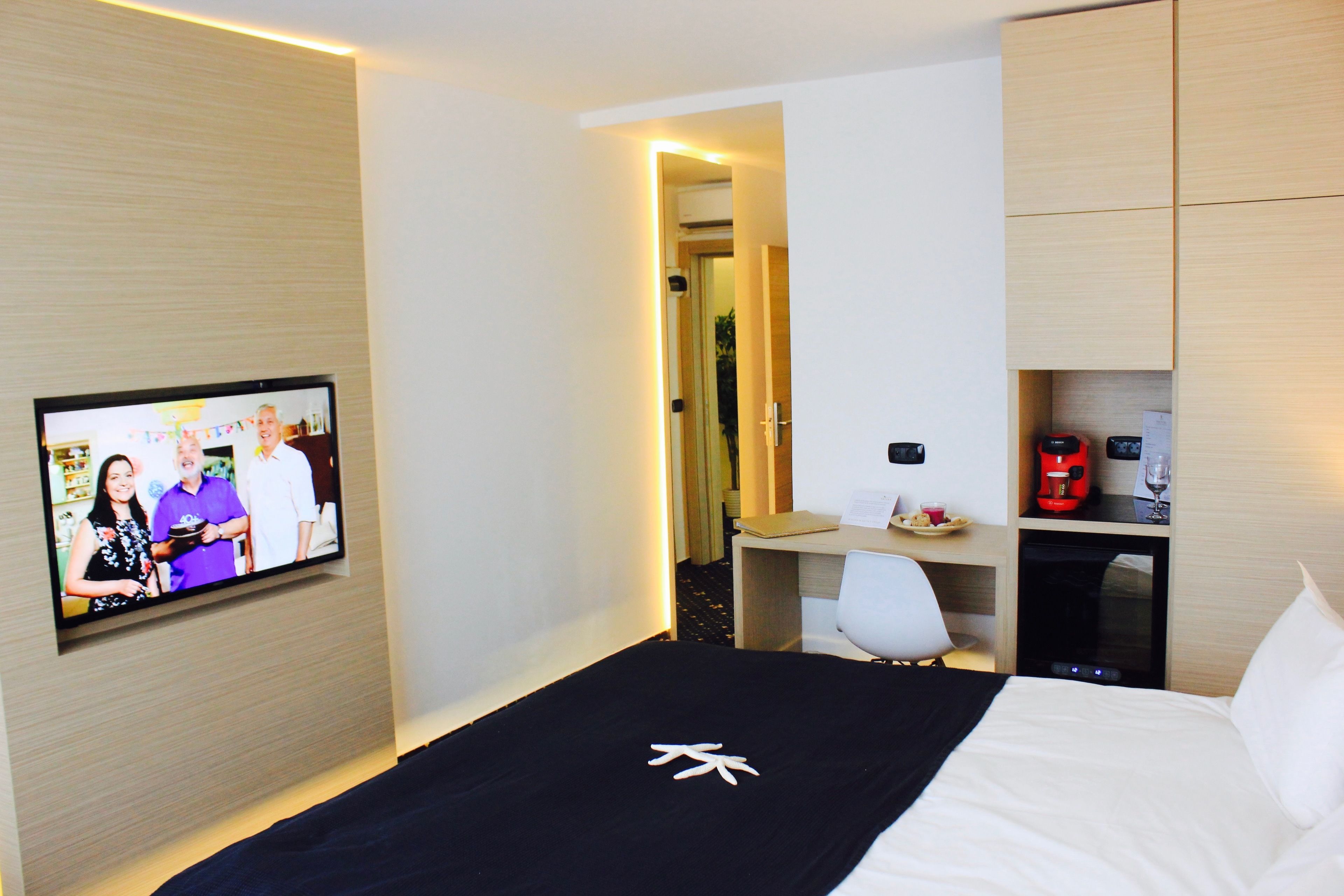 A hotel room with a bed, a TV mounted on the wall, a desk with a chair, and a minibar.