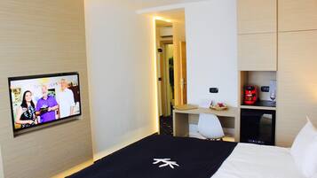 A hotel room with a bed, a TV mounted on the wall, a desk with a chair, and a minibar.