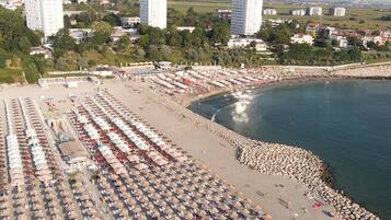 Una playa con filas de tumbonas y sombrillas, un litoral rocoso y edificios al fondo.