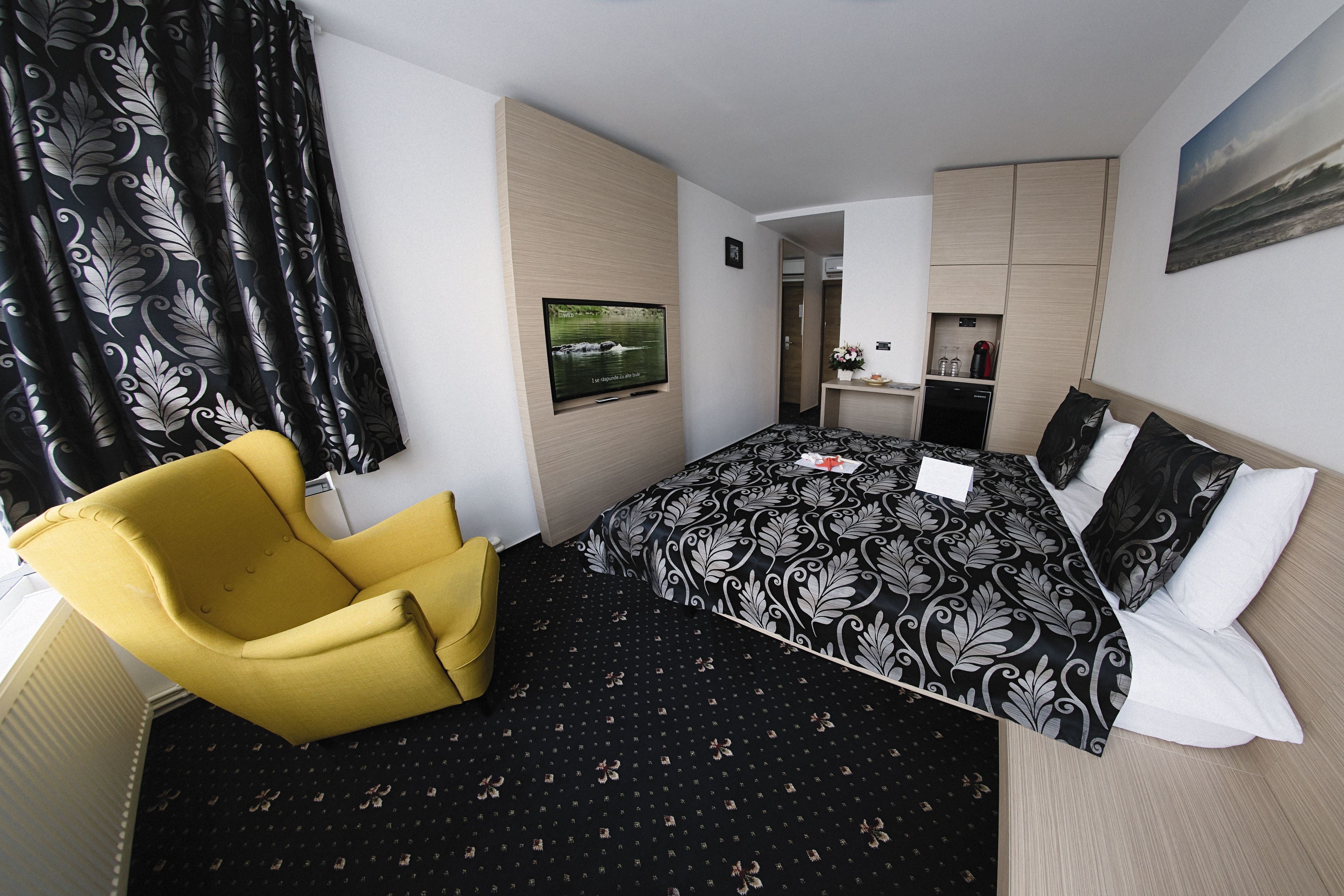 A hotel room with a bed, a yellow armchair, a flat-screen TV, a small kitchenette, and a bathroom visible through an open door.