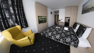 A hotel room with a bed, a yellow armchair, a flat-screen TV, a small kitchenette, and a bathroom visible through an open door.