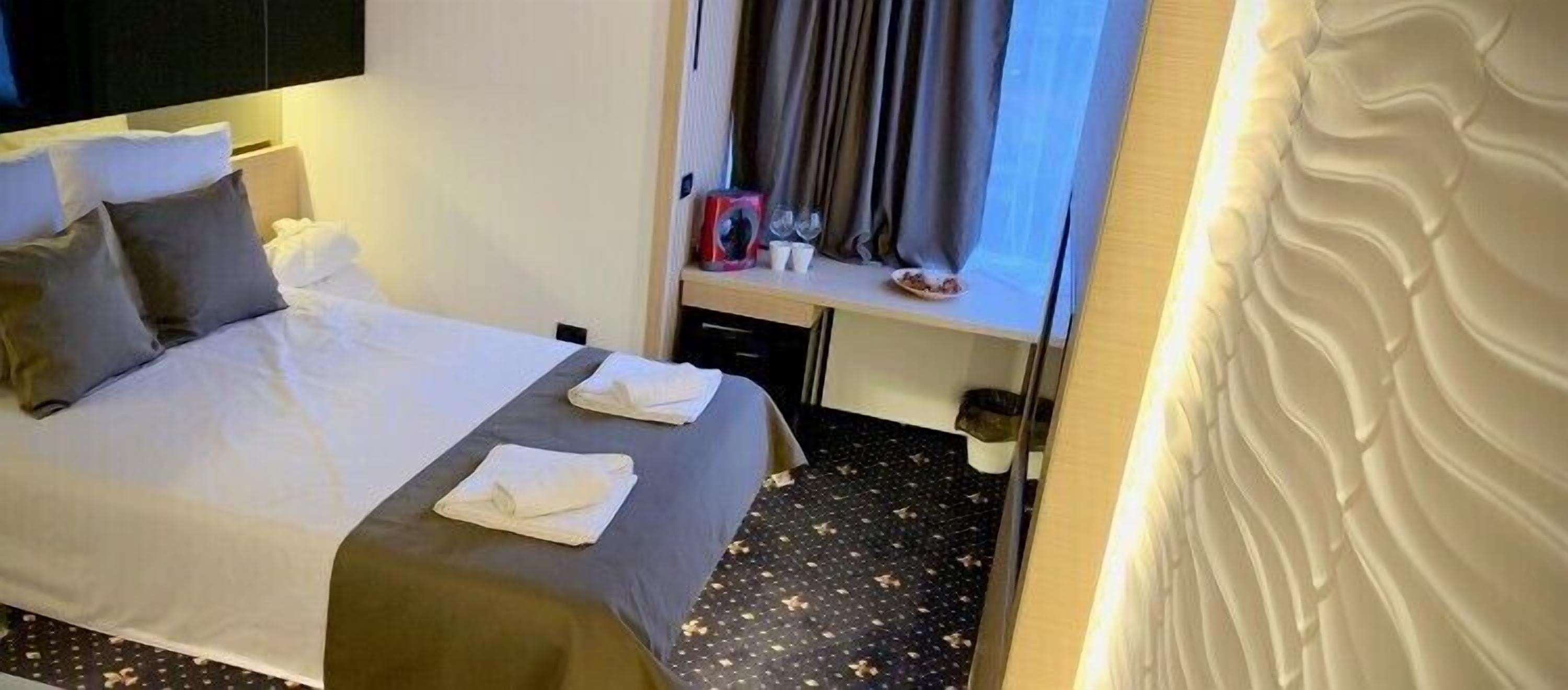 Una habitación de hotel con cama, almohadas, una mesita con una caja roja, vasos y un plato de comida.