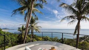 Une terrasse circulaire agrémentée d’un canapé et de coussins, offrant une vue imprenable sur la côte, avec des palmiers et la mer.