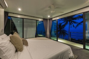 Un dormitorio moderno con una cama grande, un ventilador de techo y vistas al océano al anochecer.