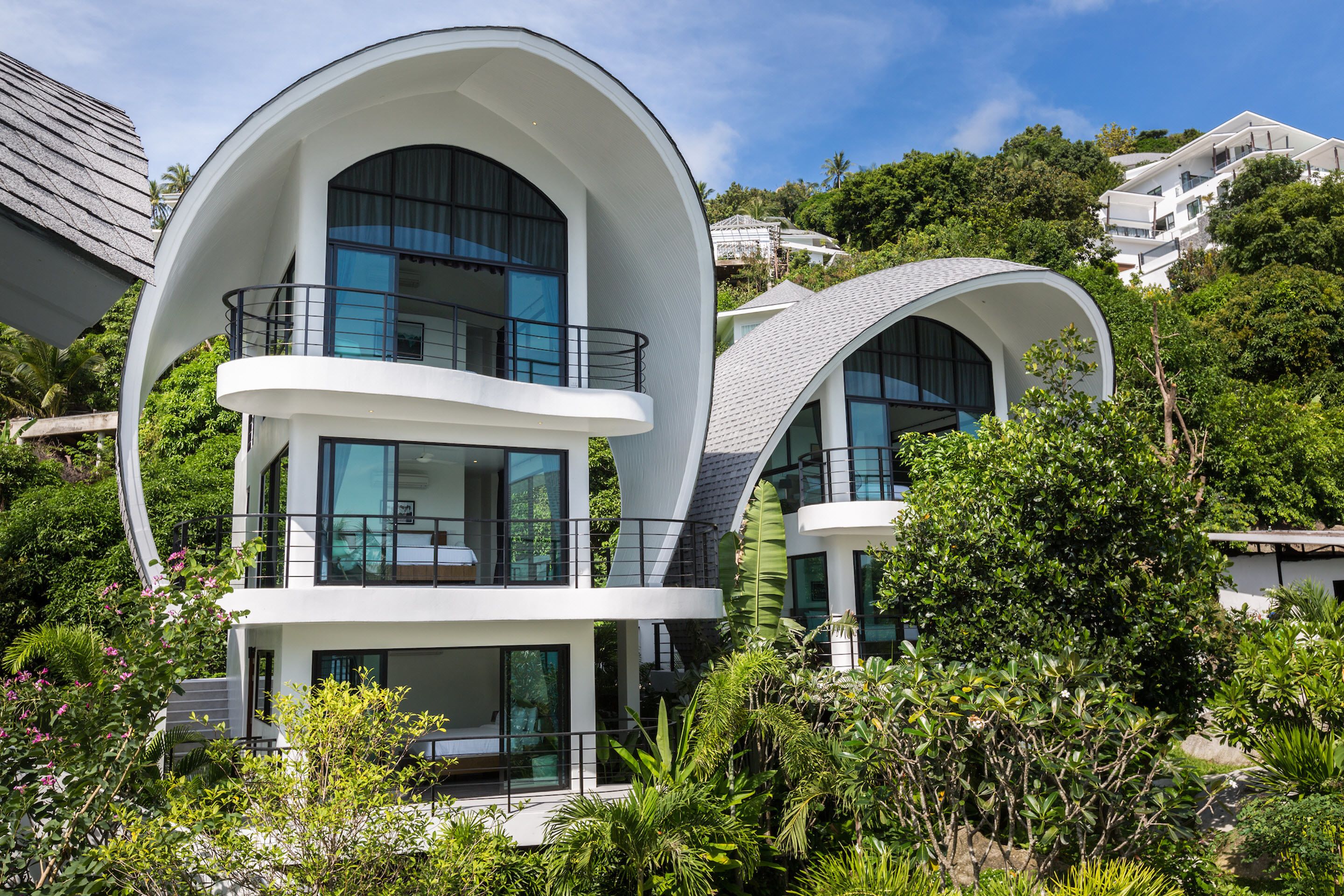 A modern, multi-level house with curved balconies and large windows, surrounded by lush greenery.