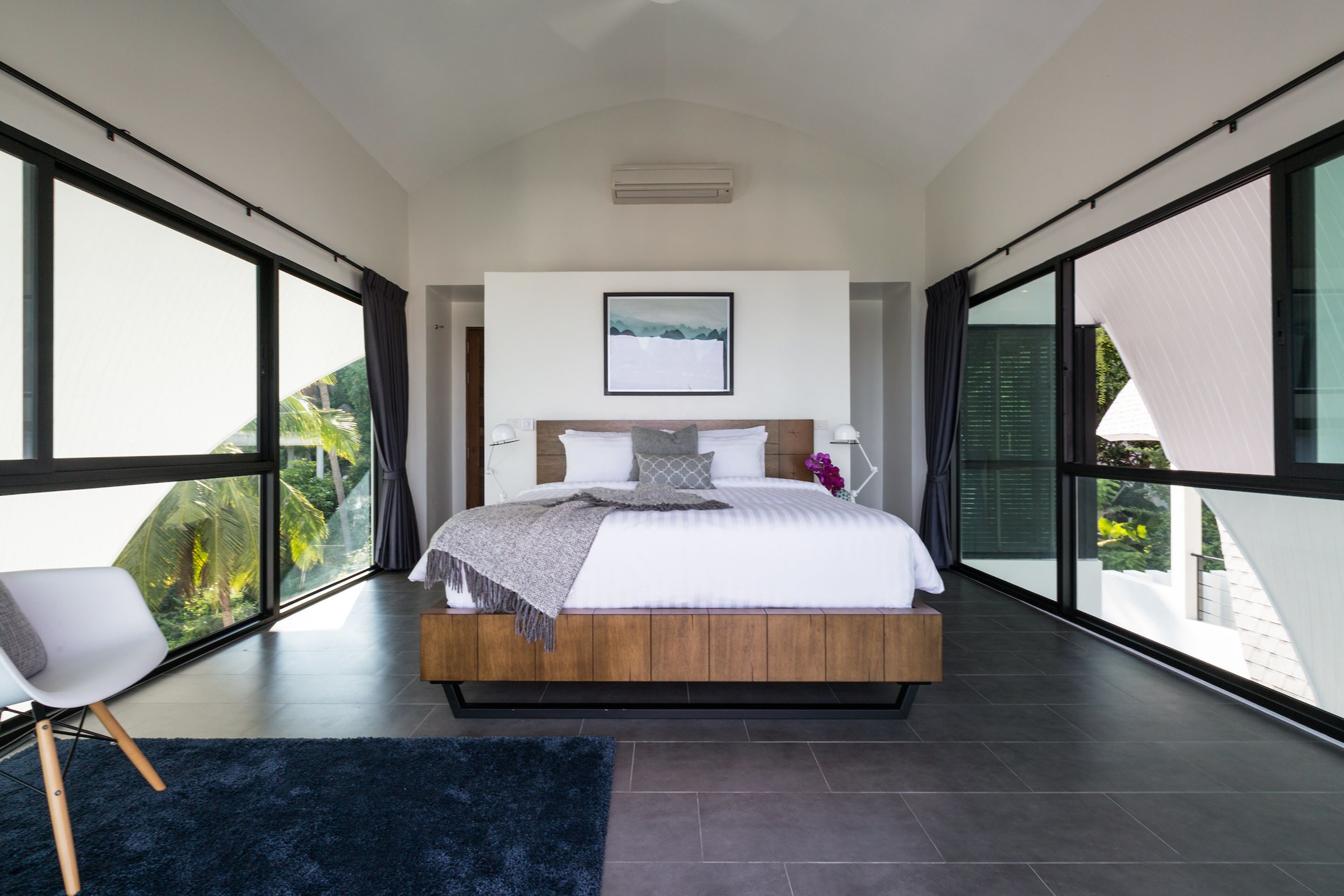A modern bedroom with a large bed, a view of greenery, and a ceiling fan.