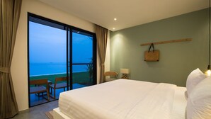 Quarto de hotel com uma cama grande, vista para o mar, varanda com cadeiras e uma bolsa pendurada.