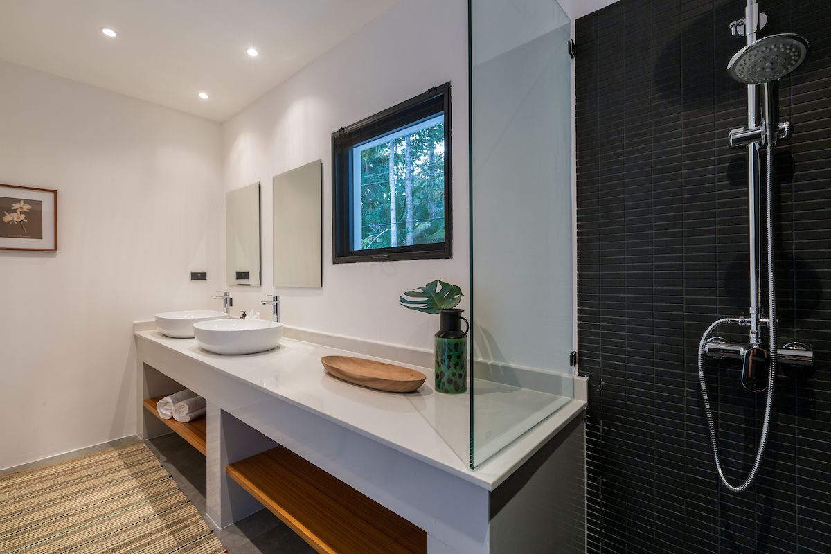 A modern bathroom with two sinks, a large mirror, a window with a view of trees, and a shower with a rain showerhead.