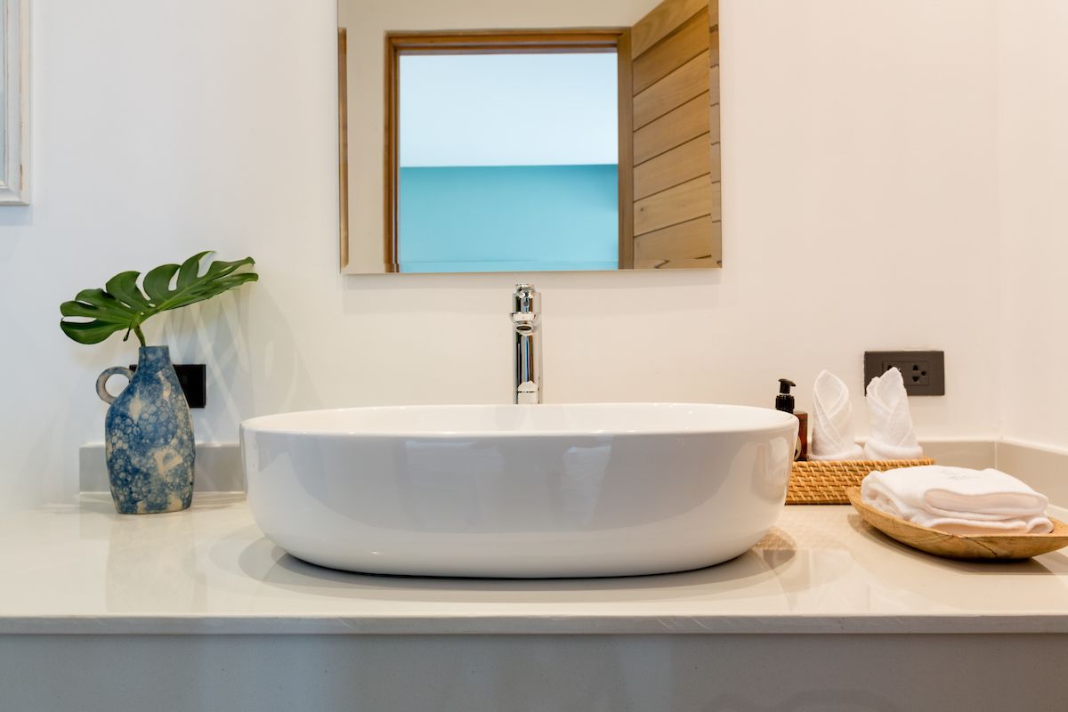 Un baño con un lavamanos grande de color blanco, un jarrón azul pequeño con una planta, una ventana de madera y un perchero para toallas.