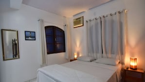 Une chambre à coucher comprenant un lit, deux tables de chevet, une petite table avec une lampe, une fenêtre avec des rideaux, un climatiseur fixé au mur et un tableau encadré.