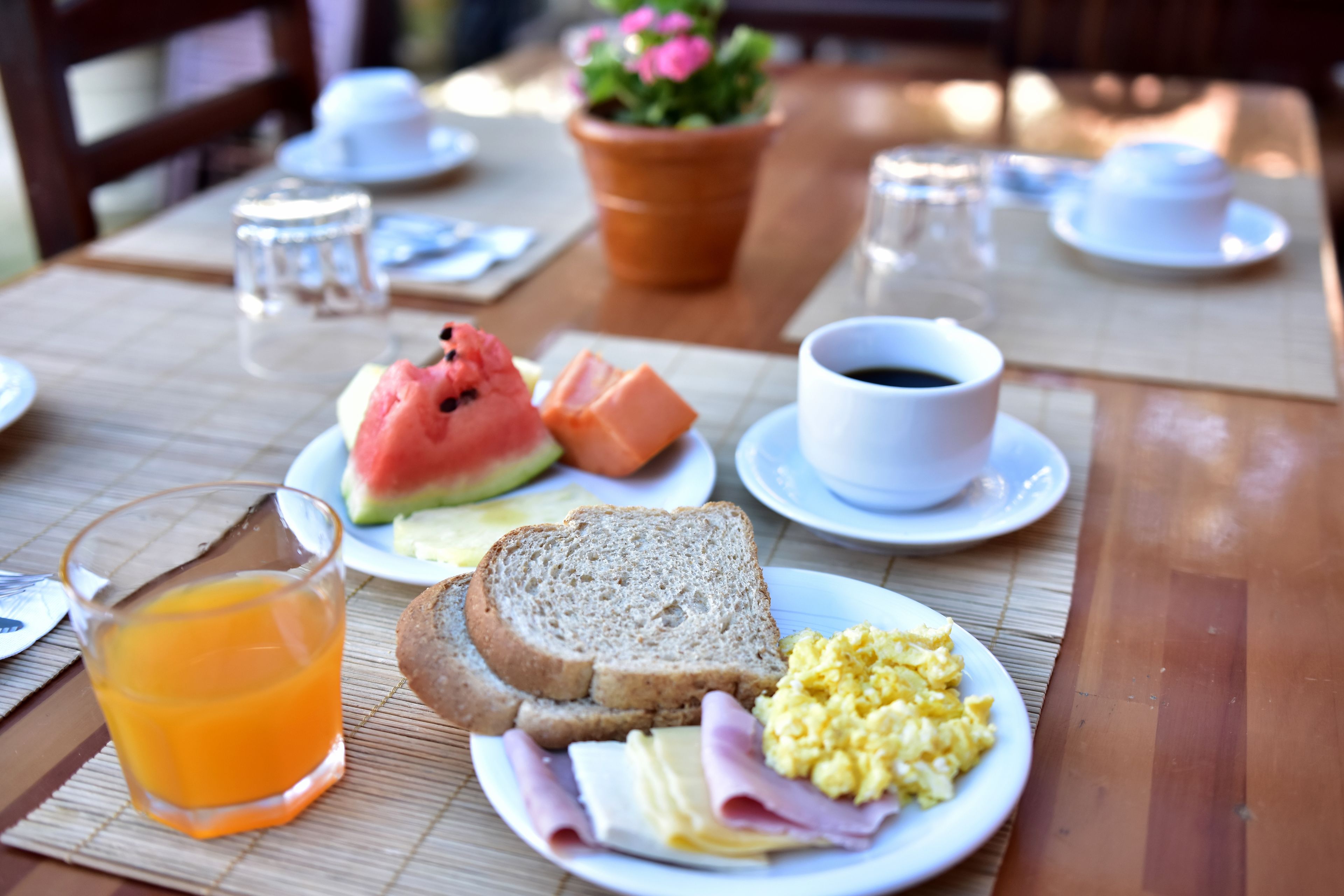 Una mesa de desayuno con tostadas, huevos, jamón y una tajada de sandía. También hay un vaso de jugo de naranja y una taza de café.