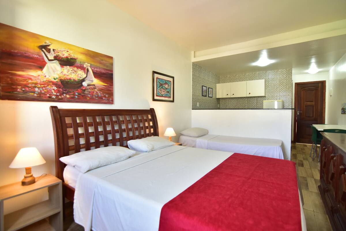 Habitación de hotel con dos camas, cabecero de madera, manta roja, un cuadro y una kitchenette al fondo.