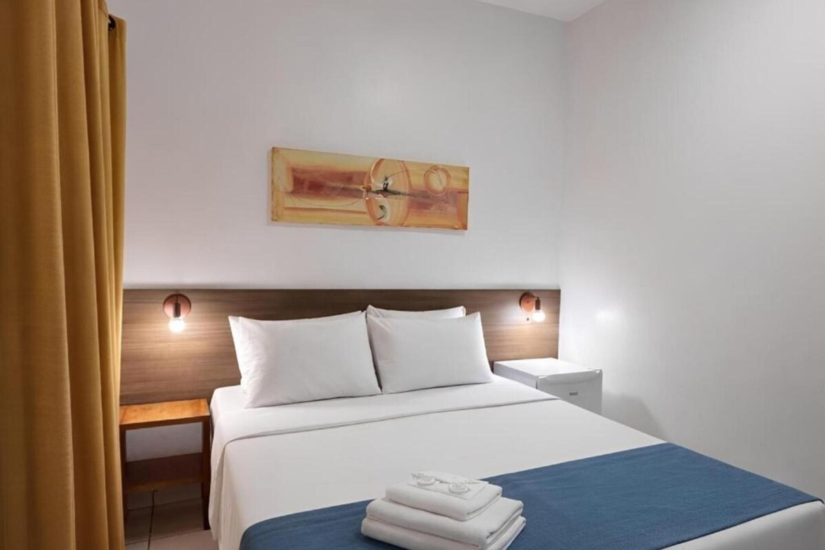 Habitación de hotel con cama, mesita de noche, lámpara de pared y un cuadro.